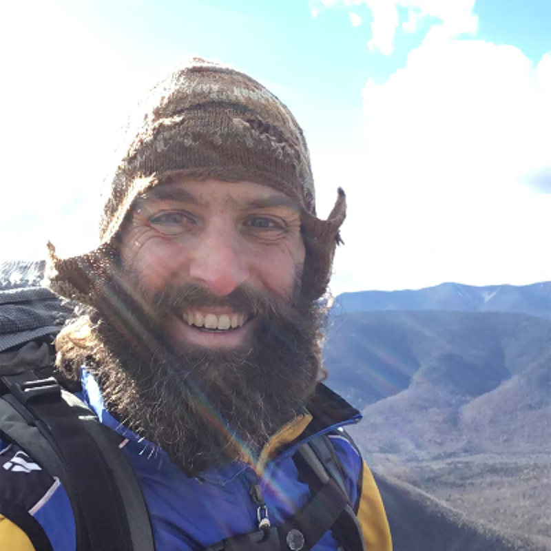The image shows a man with a long beard and a knitted hat, smiling at the camera. He appears to be outdoors, possibly hiking, with a backpack on. The background reveals a scenic view of mountains and a partly cloudy sky. The man seems happy and enjoying the outdoors.
